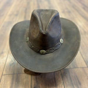 May include: A brown leather cowboy hat with a wide brim and a pinched crown. The hat features decorative metal accents around the base of the crown. The hat is made of a distressed leather material.