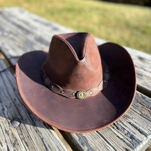 May include: A brown leather cowboy hat with a wide brim and a decorative band featuring metal accents. The hat is sitting on a weathered wooden surface, suggesting an outdoor setting. The hat's design is classic and rugged.