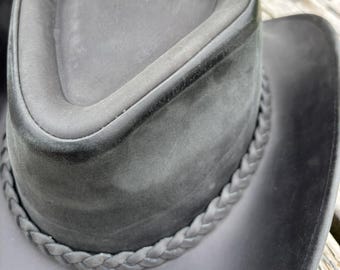 May include: A dark gray cowboy hat with a braided band around the brim. The hat has a rounded crown and a wide, curved brim. The hat appears to be made of leather or a similar material.