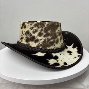 May include: Cowboy hat with a spotted pattern in brown and cream on the crown. The brim is black with a matching braided band around the base of the crown. A classic western style hat.