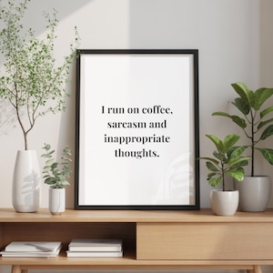 Op de afbeelding: Een ingelijste print met de tekst "I run on coffee, sarcasm and inappropriate thoughts." De zwart omlijste print wordt weergegeven op een houten console, samen met decoratieve planten in witte en grijze potten en vazen.