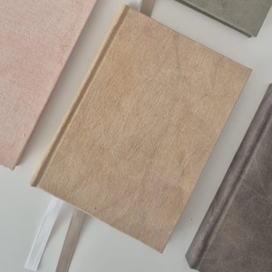 Puede incluir: Cinco cuadernos de diferentes colores con marcadores de cinta blanca. Los cuadernos están hechos de papel texturizado y tienen un aspecto rústico.