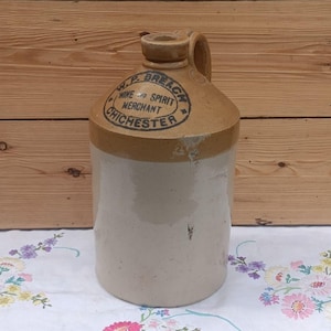 Puede incluir: Una jarra cilíndrica de gres con una sección superior bronceada y un cuerpo inferior de color crema. La jarra cuenta con un asa y texto impreso en negro que dice W.P. BREACH WINE AND SPIRIT MERCHANT CHICHESTER. La superficie muestra desgaste.