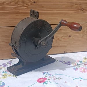 May include: A vintage cast iron kitchen tool with a circular body and a dark grey finish. It features a side-mounted hand crank with a smooth, dark brown wooden handle. The metal casing has embossed lettering and a sturdy base for stability.
