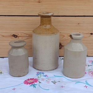 Könnte beinhalten: Drei zylindrische Steinzeugflaschen mit engem Hals und ausgestelltem Rand. Die mittlere Flasche ist höher, hat einen beigefarbenen Hals und einen cremeweißen, gesprenkelten Körper. Die beiden kleineren Flaschen sind hellgrau und gesprenkelt.