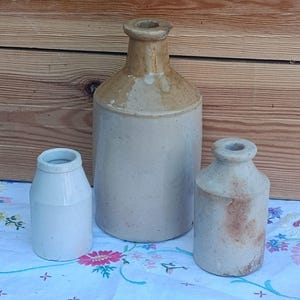 Könnte beinhalten: Drei antike Steinzeugflaschen in verschiedenen Größen. Die größte Flasche ist beige und hellbraun, mit rundem Körper und schmalem Hals. Die anderen beiden Flaschen sind kleiner, mit ähnlicher Form und Farbe. Sie stehen auf einer Tischdecke mit Blumenmuster.