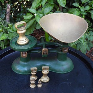 Puede incluir: Una balanza de estilo antiguo con base y estructura de hierro fundido en color verde oscuro y un gran plato de pesaje de latón. Incluye un juego de cinco pesas de latón de distintos tamaños, cada una con asa, situadas sobre la base y la plataforma.