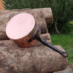 May include: A copper pot with a black handle. The pot has a brass rim and is resting on a pile of logs.
