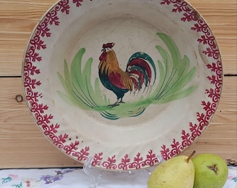 May include: A vintage, off-white ceramic plate with a hand-painted rooster in the center, surrounded by green brushstrokes. The rim features a red floral design. Two pears rest in front of the plate.
