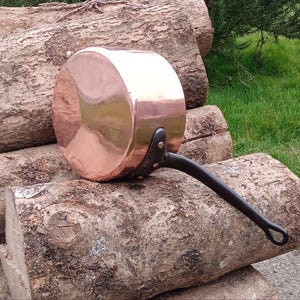 May include: A polished copper saucepan with a smooth, reflective circular base and a long, dark, curved metal handle attached with two visible rivets. The pan features a cylindrical body and a flat bottom, showcasing a metallic finish.