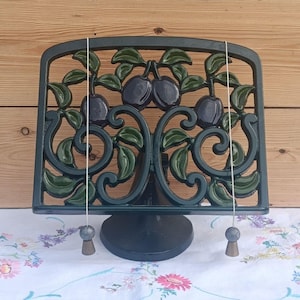 May include: A dark green metal cookbook stand featuring an openwork design of dark purple plums and green leaves. The stand includes two thin metal arms with small weighted ends hanging down to hold pages open, supported by a circular base.