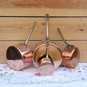 May include: Three copper pans with silver-coloured handles are arranged on a floral patterned tablecloth. The pans vary in size, with a small pan nested inside a larger one. The background is a wooden wall.
