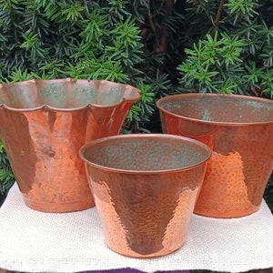 Puede incluir: Tres macetas de cobre para plantas, una con borde festoneado, sobre una superficie blanca texturizada. Las macetas están todas martilladas y tienen un aspecto rústico y envejecido.