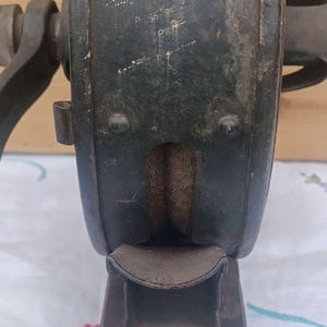 May include: A vintage dark green metal hand-cranked tool featuring a central circular housing with a visible internal grinding wheel, a curved metal discharge chute at the bottom, and a sturdy rectangular base with mounting holes for secure attachment.
