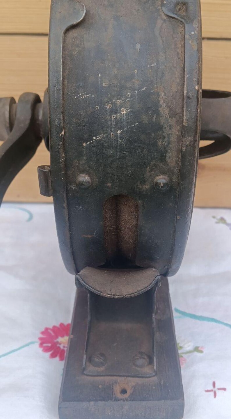 May include: A vintage dark green metal hand-cranked tool featuring a central circular housing with a visible internal grinding wheel, a curved metal discharge chute at the bottom, and a sturdy rectangular base with mounting holes for secure attachment.