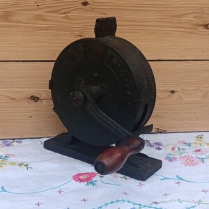May include: A vintage cast iron hand-cranked tool with a circular body and a dark, weathered finish. It features a central rotating arm with a smooth, dark brown wooden handle attached to a rectangular base, showing faint embossed lettering.