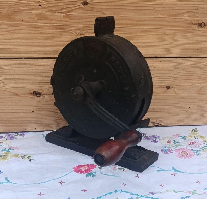 May include: A vintage cast iron hand-cranked tool with a circular body and a dark, weathered finish. It features a central rotating arm with a smooth, dark brown wooden handle attached to a rectangular base, showing faint embossed lettering.