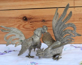 May include: Two metallic rooster figurines crafted with intricate etched detailing on the feathers and bodies. One rooster stands upright with a prominent tail, while the other is positioned in a lower, forward-leaning stance with wings spread.