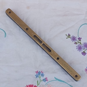 May include: A vintage brass level with two bubble vials. The level has a rectangular shape with rounded ends and is marked with text. The level is lying on a white floral patterned cloth.