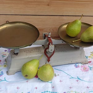Peut inclure: Une ancienne balance de cuisine avec deux plateaux en laiton, un bras rouge et blanc, et une base blanche. Trois poires vertes sont sur la balance et une nappe fleurie. La balance est contre un fond en bois.