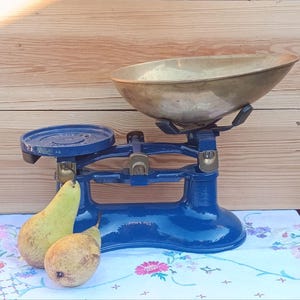 May include: A classic blue and brass scale featuring a brass bowl and a round platform. Two pears are placed on a floral cloth in front of the scale. The background is wooden.