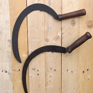 Peut inclure: Deux faucilles à main dotées de lames métalliques noires incurvées et de manches en bois brun foncé. Chaque outil présente une lame tranchante en forme de croissant fixée à une poignée cylindrique, conçue pour les travaux de récolte manuelle.