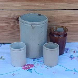 Pode incluir: Uma coleção de potes e jarras de grés antigos. O grupo inclui um grande pote cilíndrico cinzento, dois potes menores creme e um pote vidrado castanho. Os artigos estão dispostos sobre uma toalha de mesa com padrão floral.