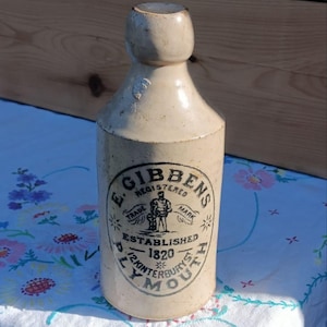 Puede incluir: Botella antigua de gres con acabado color crema. La botella presenta una etiqueta circular con el texto "E. Gibbens Registered Trade Mark Established 1820 Plymouth". La botella se exhibe sobre un mantel floral.