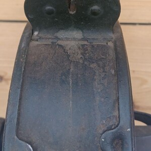 May include: A dark, weathered metal component featuring a curved, plate-like structure with a distinct notched top section. The surface displays signs of oxidation, rust, and wear, with visible rivets securing the upper bracket to the main body.