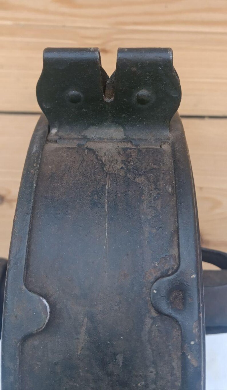 May include: A dark, weathered metal component featuring a curved, plate-like structure with a distinct notched top section. The surface displays signs of oxidation, rust, and wear, with visible rivets securing the upper bracket to the main body.