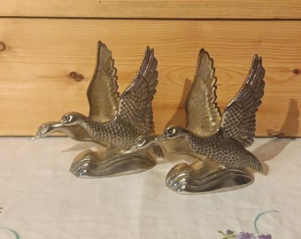 May include: Two silver-colored duck figurines in flight, with outstretched wings and detailed feather textures. The ducks are mounted on a wave-like base, creating a dynamic and decorative display. The background is a light-colored surface and a wooden wall.