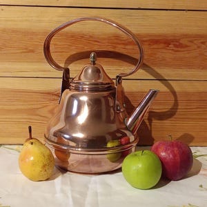 May include: A polished copper kettle with a curved handle and spout. A yellow pear, a green apple, and a red apple sit beside the kettle. The background is a wooden wall.