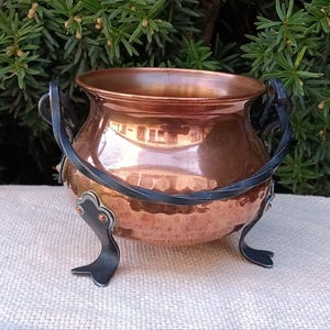 May include: A small copper cauldron with black iron handles and three legs. The cauldron has a hammered texture and is a decorative piece.