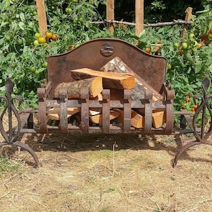 May include: A rustic, dark brown cast iron fireplace grate featuring a curved backplate with a small circular emblem. The grate has vertical bars and two decorative fleur-de-lis style side supports, currently holding several pieces of cut firewood.