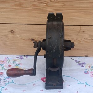 May include: A vintage cast iron hand-cranked tool featuring a dark, weathered metal body with a central circular housing and a sturdy base. A wooden handle with a dark brown finish is attached to the side crank arm for manual operation.