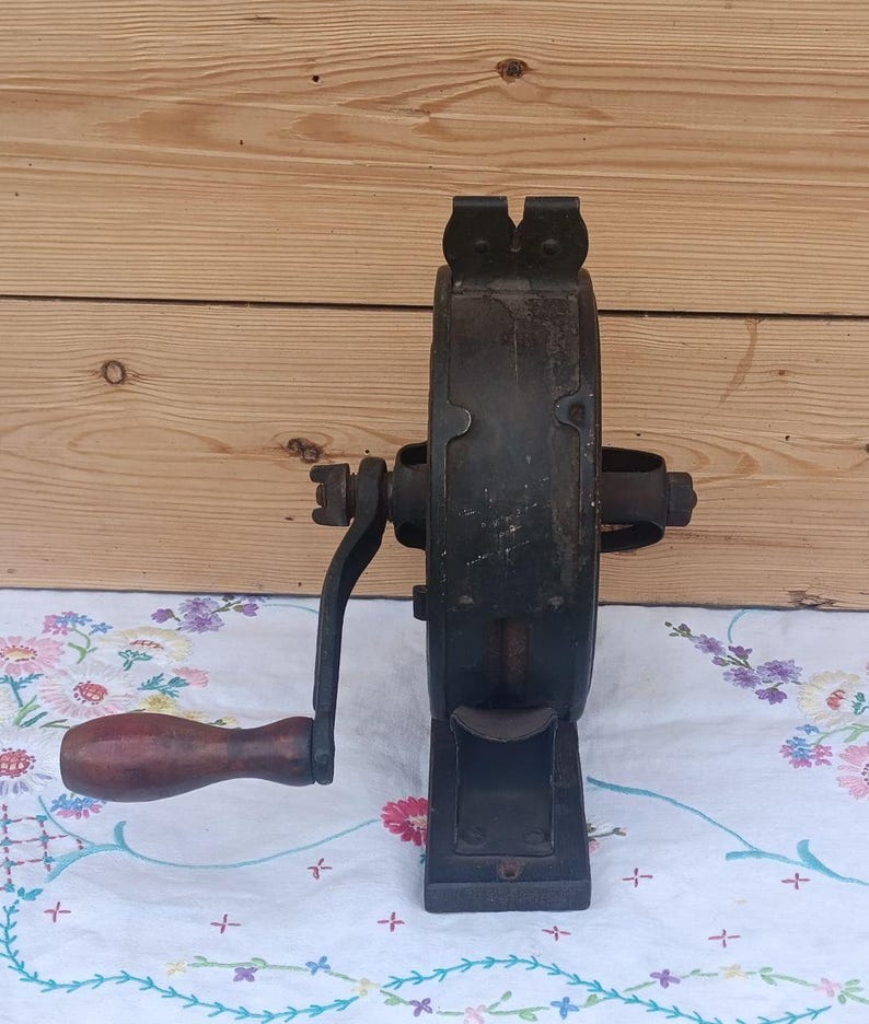May include: A vintage cast iron hand-cranked tool featuring a dark, weathered metal body with a central circular housing and a sturdy base. A wooden handle with a dark brown finish is attached to the side crank arm for manual operation.