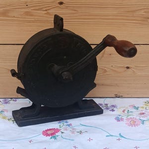 May include: A vintage black cast iron hand-cranked food mill featuring a circular body with embossed lettering, a sturdy rectangular base, and a smooth brown wooden handle attached to a metal crank arm for manual operation of the internal mechanism.