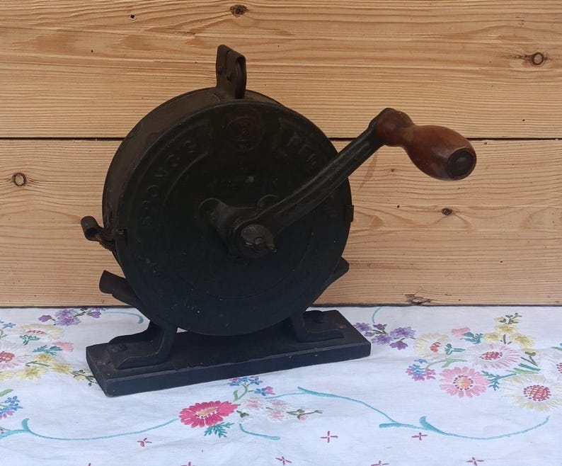 May include: A vintage black cast iron hand-cranked food mill featuring a circular body with embossed lettering, a sturdy rectangular base, and a smooth brown wooden handle attached to a metal crank arm for manual operation of the internal mechanism.