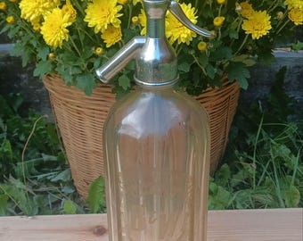 Pode incluir: Garrafa de seltzer de vidro vintage com tampa e alavanca cromadas. A garrafa transparente tem um design canelado. O fundo apresenta uma cesta de vime cheia de flores amarelas.