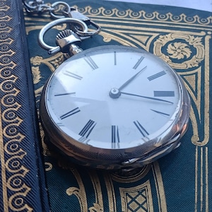 May include: A silver-toned pocket watch with a white face, black Roman numeral hour markers, and thin black hands. The watch features a textured metallic edge and is attached to a silver-toned metal chain link, showcasing a classic circular design.