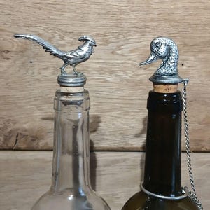以下が含まれることがあります： 鳥のデザインが施された2つの装飾的なボトルストッパー。1つはキジ、もう1つはアヒルのデザインです。どちらも銀色の金属製で、コルクの上にあります。アヒルのストッパーにはチェーンが付いています。ボトルは透明と茶色です。
