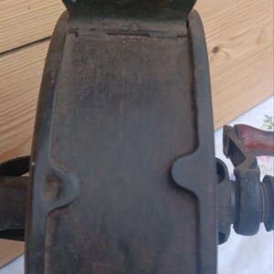 May include: A dark, weathered cast iron grinding wheel featuring a thick, circular metal frame with a prominent top bracket and a side-mounted handle with a wooden grip. The surface shows signs of oxidation and age, with a sturdy, industrial design.