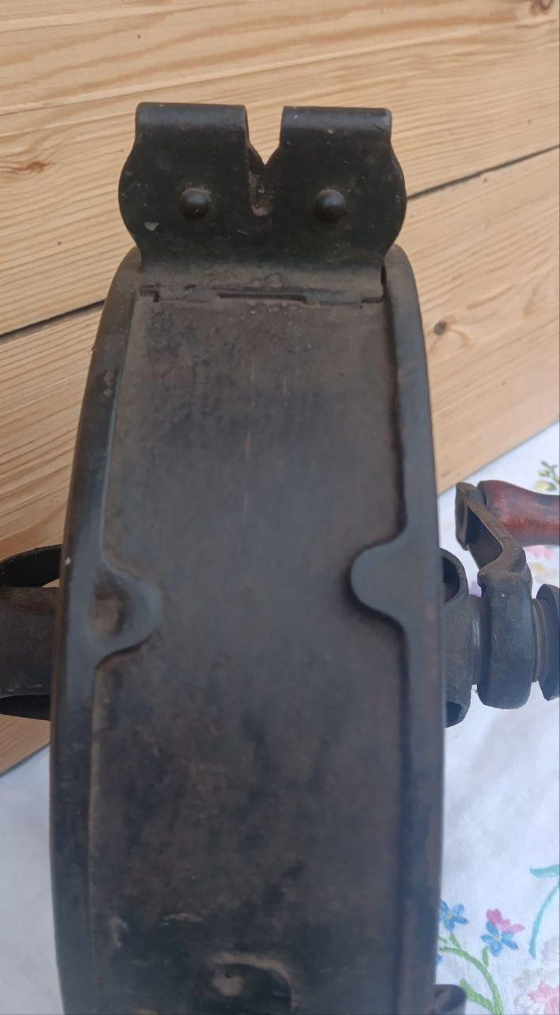 May include: A dark, weathered cast iron grinding wheel featuring a thick, circular metal frame with a prominent top bracket and a side-mounted handle with a wooden grip. The surface shows signs of oxidation and age, with a sturdy, industrial design.