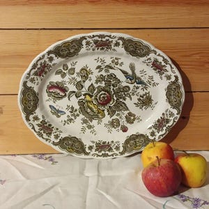 Puede incluir: Una fuente ovalada con un estampado floral y de pájaros en tonos verdes, marrones, amarillos y rojos. La fuente tiene un borde festoneado. Tres manzanas están frente a la fuente, sobre un mantel blanco.