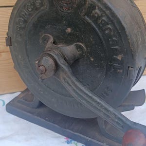 May include: A vintage black cast iron Spong's food mill featuring a circular housing with embossed lettering, a central hand-crank mechanism, and a wooden handle. The device is mounted on a rectangular base and displays signs of age and wear.