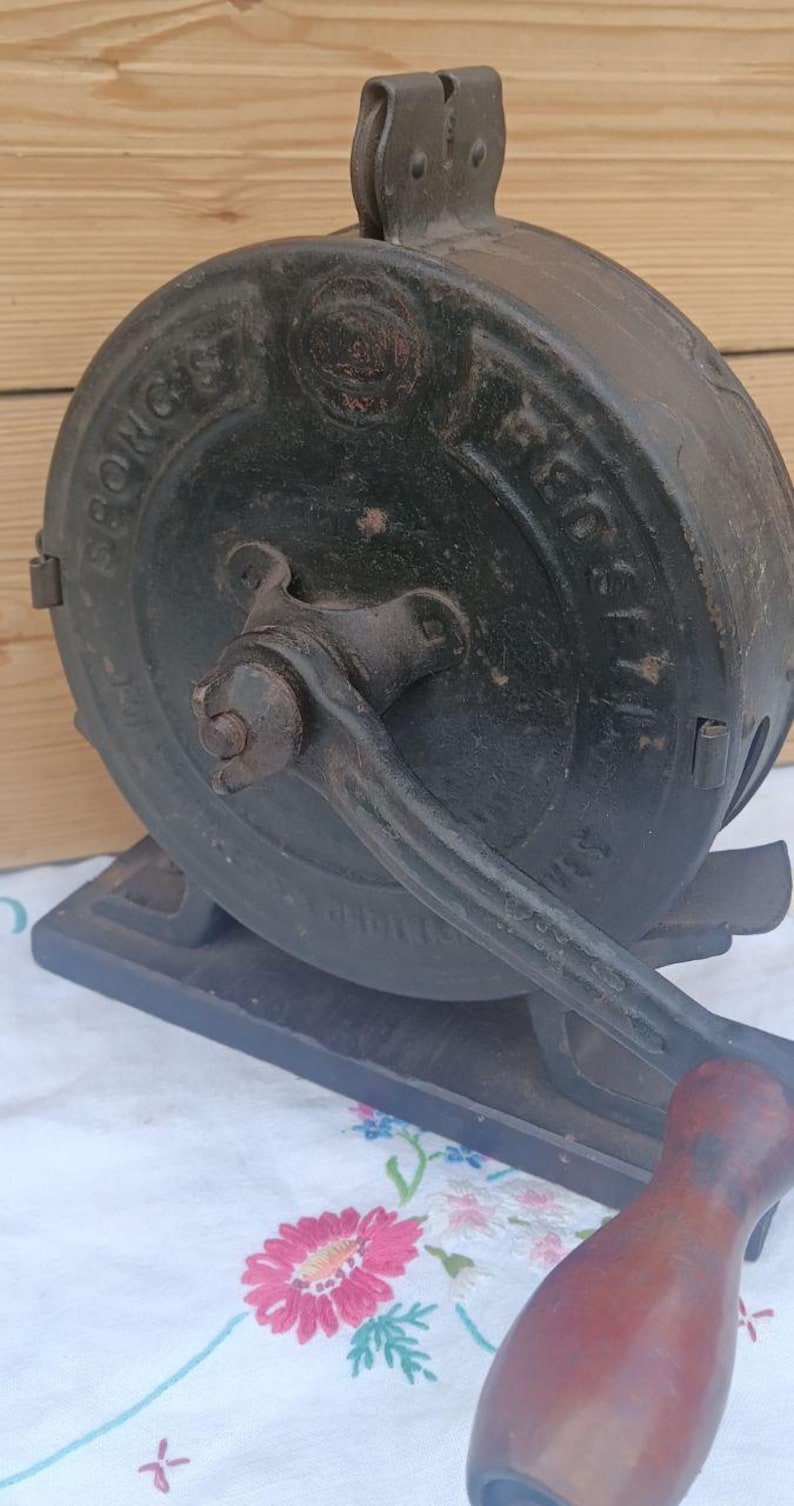 May include: A vintage black cast iron Spong's food mill featuring a circular housing with embossed lettering, a central hand-crank mechanism, and a wooden handle. The device is mounted on a rectangular base and displays signs of age and wear.