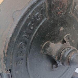 May include: A vintage black cast iron kitchen tool with a circular body and a central handle. The word "SPONG'S" is embossed in raised lettering on the side of the circular housing. The metal surface shows signs of age, wear, and texture.