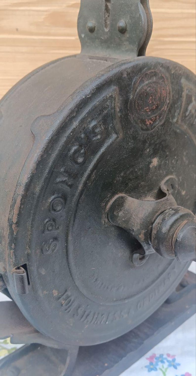 May include: A vintage black cast iron kitchen tool with a circular body and a central handle. The word "SPONG'S" is embossed in raised lettering on the side of the circular housing. The metal surface shows signs of age, wear, and texture.