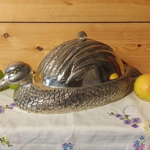 Può includere: Un piatto da portata argentato a forma di anatra con un design dettagliato a piume sul coperchio e sul corpo. Il piatto è posto su una tovaglia floreale, con una mela verde e una pera gialla su entrambi i lati.