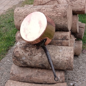 May include: A copper pot with a long black handle. The pot is sitting on a pile of logs.